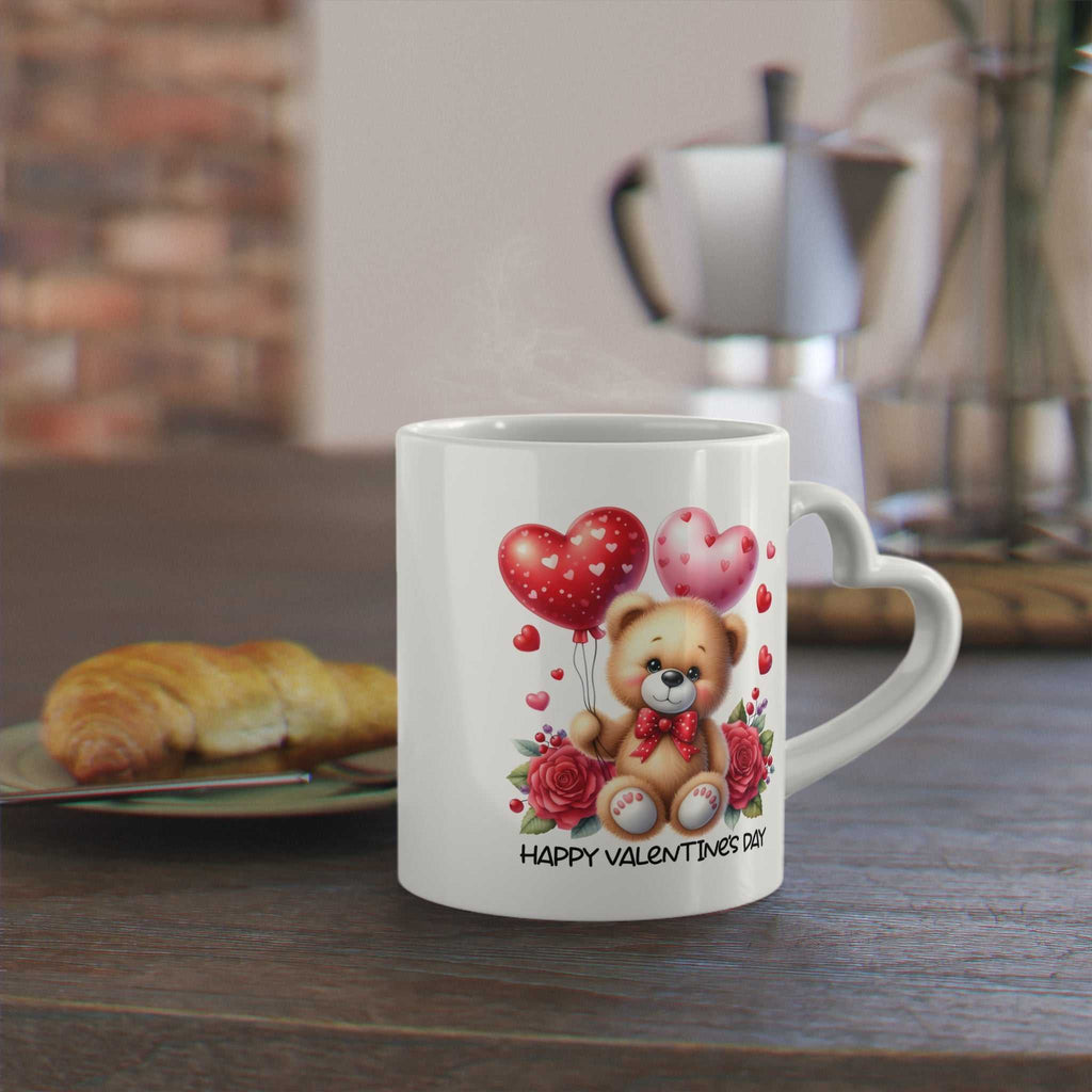 Cute Valentines Day mug with heart-shaped handle featuring a teddy bear and heart balloons design on a table.