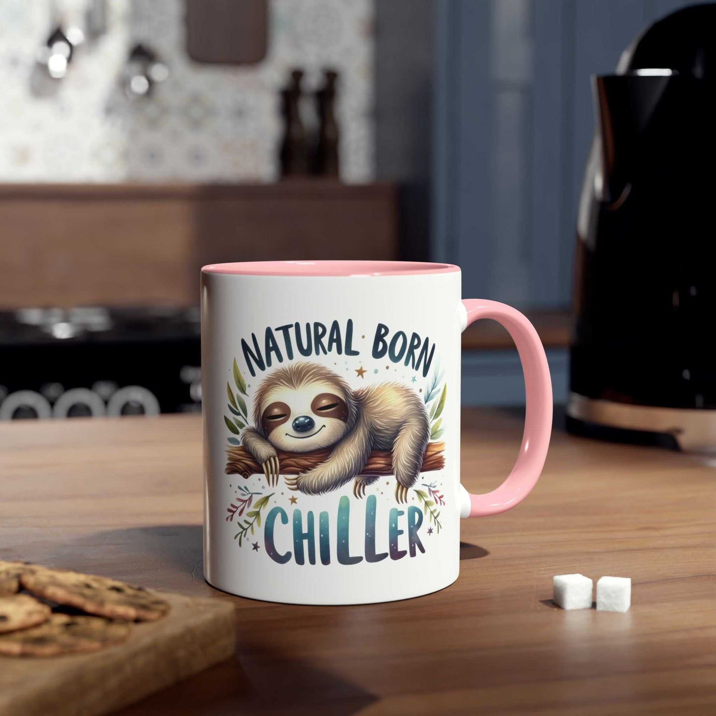 Cute sloth coffee mug with "Natural Born Chiller" design on wooden table in cozy kitchen setting.