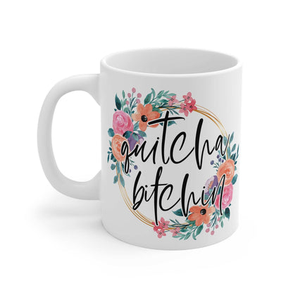 White ceramic mug with "quitcha bitchin" text surrounded by colorful floral design, glossy finish, 11oz capacity.