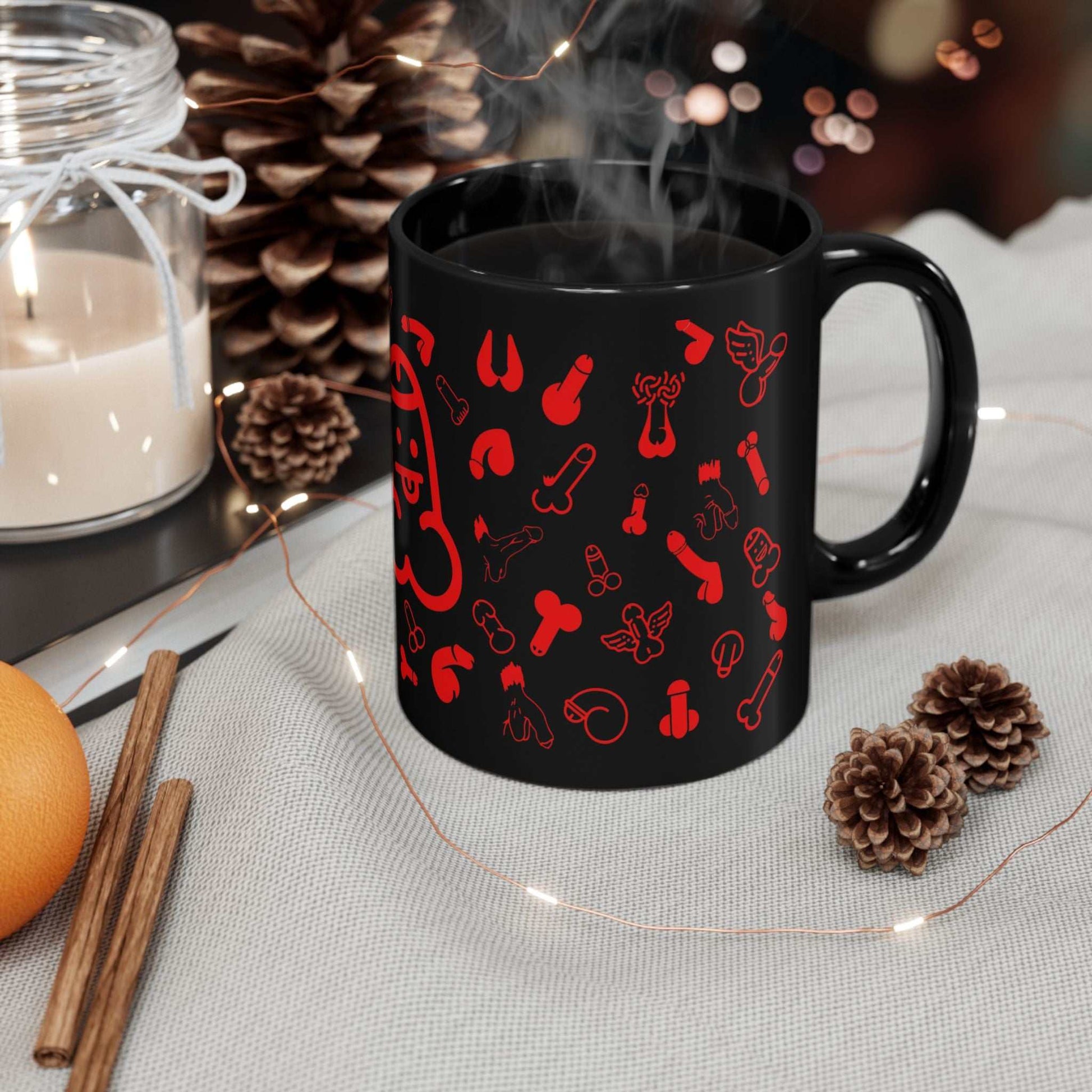 Black ceramic mug with playful red willie print, resting on a festive table setup with pinecones and a candle.