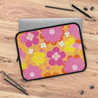 Retro floral laptop sleeve in vibrant pink, yellow, and orange flowers with black neoprene edges, protecting a laptop on wooden surface.