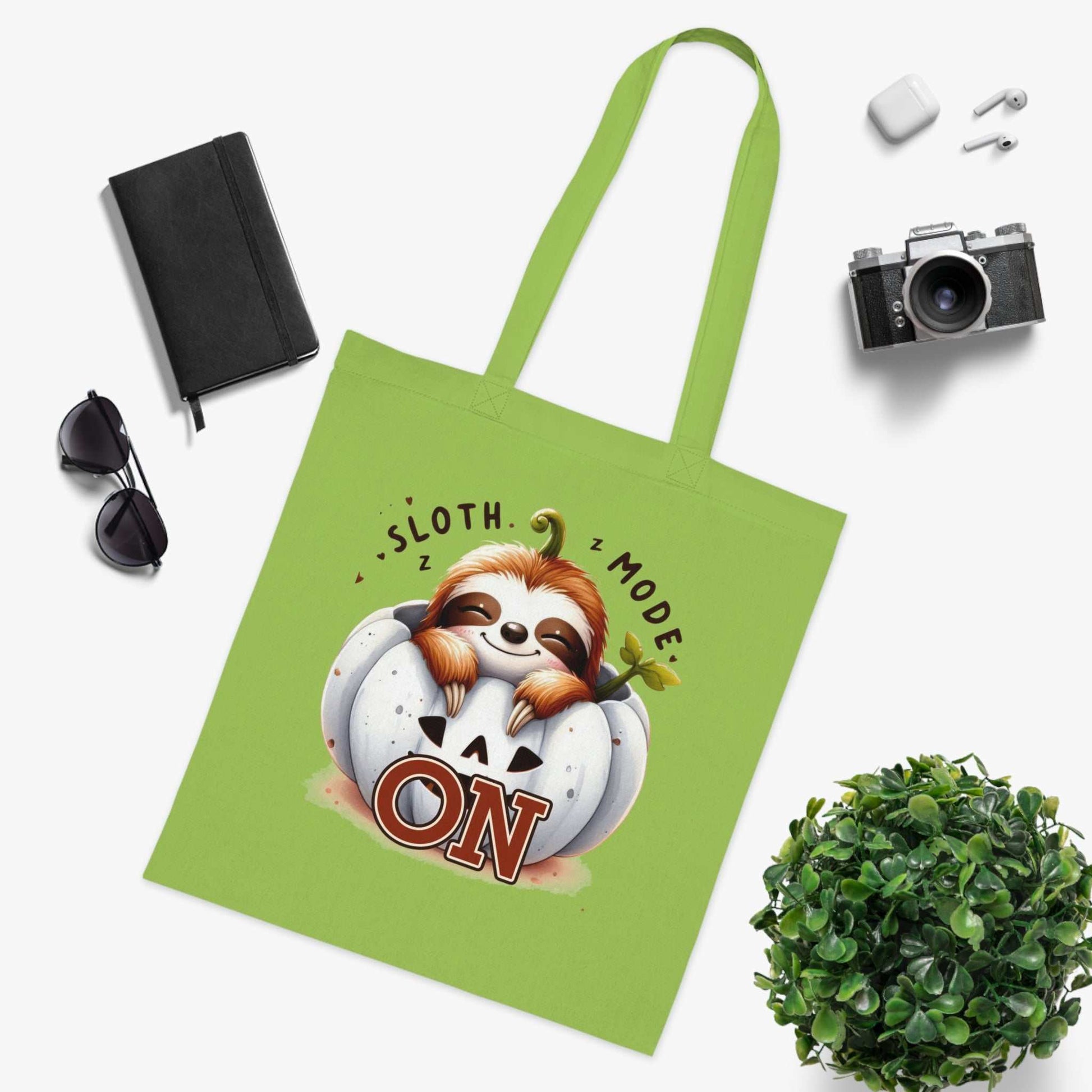 Cute sloth tote bag with "Sloth Mode On" design, green cotton fabric, surrounded by sunglasses, notebook, camera, and plant.