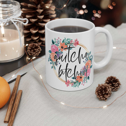 White ceramic coffee mug with "Quitcha Bitchin" text surrounded by colorful floral design, glossy finish, filled with coffee on a table with cozy decor.