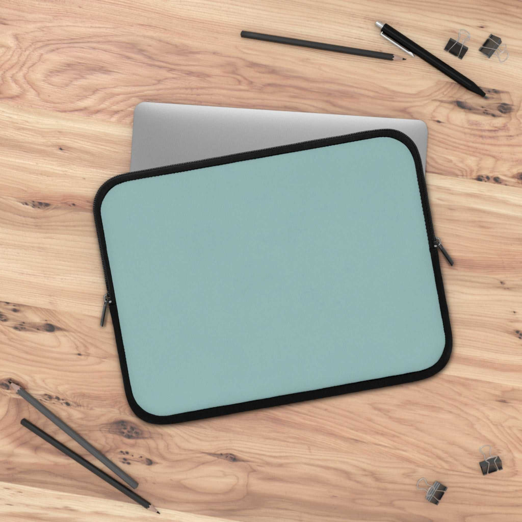 teal green neoprene laptop sleeve with black trim on wooden desk with pencils and clips