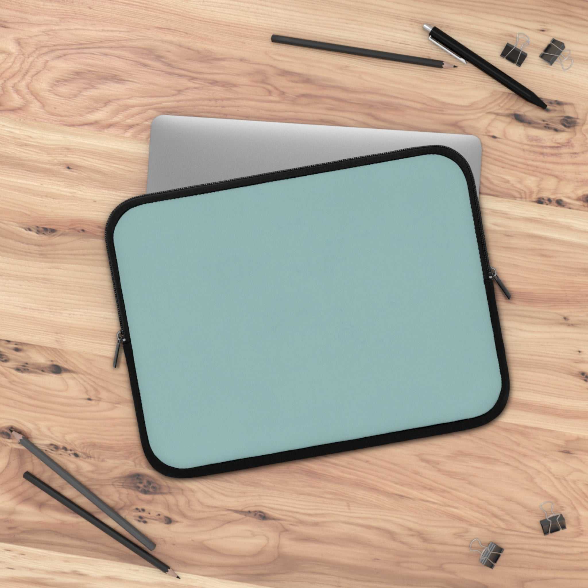 teal green neoprene laptop sleeve with black trim on wooden desk with pencils and clips