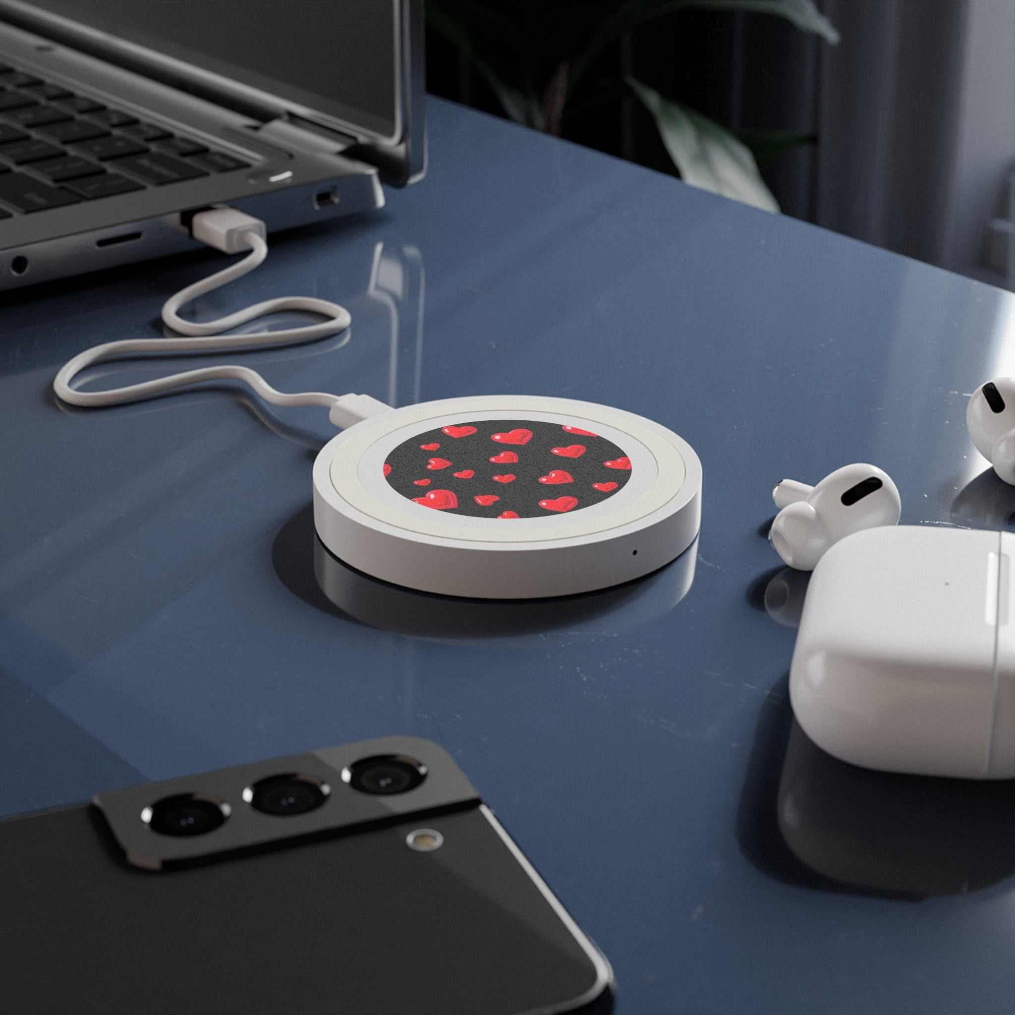 Compact black wireless charging pad with red heart design, 5W power, and USB cable on desk with smartphone and earbuds.