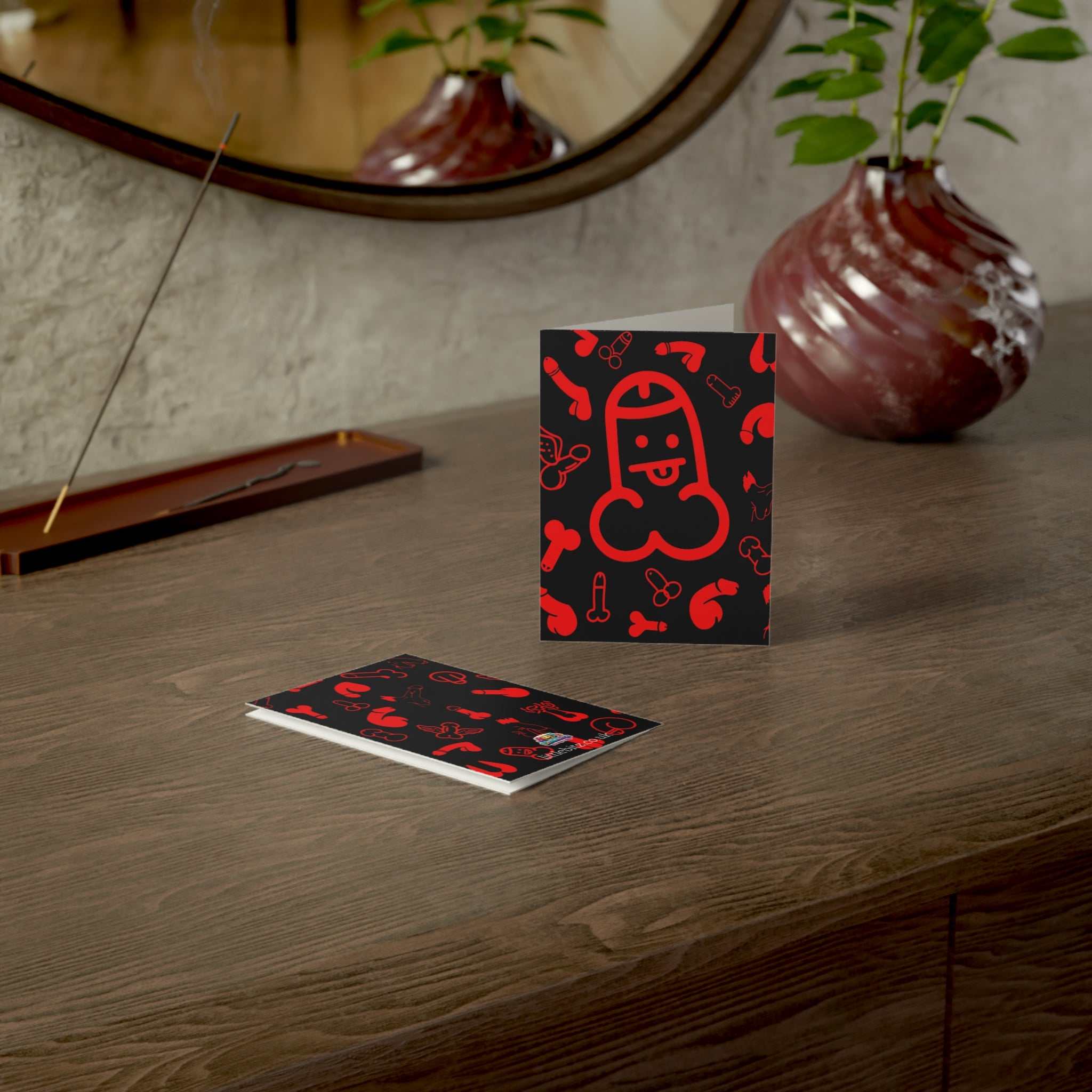 Cheeky Rude Red Willie Greeting Card on a wooden table with a plant and mirror in the background.